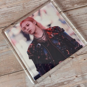 May include: A square glass coaster featuring a printed image of a person with light pink hair, wearing a dark jacket adorned with multiple silver buckles and layered silver chain necklaces. The coaster has a clear, thick border and a smooth finish.