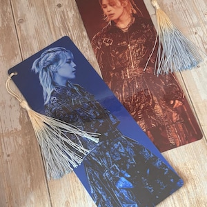May include: Two rectangular bookmarks featuring a person with light hair in a dark jacket. One bookmark has a deep blue color scheme, and the other has a warm reddish-brown tone. Each bookmark includes a hole at the top with a decorative tassel.