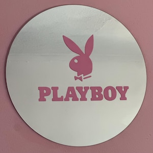 May include: A circular mirror featuring a pink Playboy bunny logo and the word PLAYBOY in pink lettering centered on the reflective surface. The design is applied directly to the glass, creating a clear reflection of the surrounding environment.