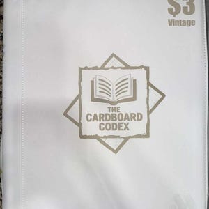 以下が含まれることがあります： 「THE CARDBOARD CODEX」の文字と開いた本のグラフィックが描かれた白いバインダー。右上には「$3 Vintage」の文字。バインダーは滑らかな素材でできており、ステッチされたエッジがあります。