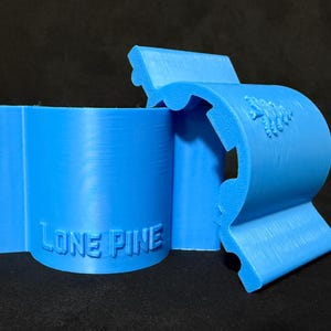 Könnte beinhalten: Hellblaues 3D-gedrucktes Objekt mit dem geprägten Text "LONE PINE". Das Objekt scheint eine Klammer oder Halterung zu sein, mit einem geschwungenen Design und einem kleinen geprägten Emblem. Schwarzer Hintergrund.