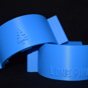 Op de afbeelding: Twee helderblauwe, gebogen objecten met rechthoekige verlengingen. Eén heeft een reliëfdennenboom, de andere leest "LONE PINE" in hoofdletters. De objecten zijn waarschijnlijk decoratief of functioneel.