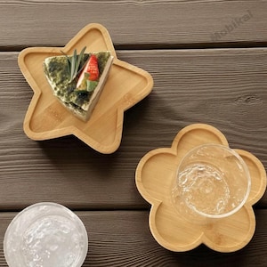 May include: Wooden star-shaped and flower-shaped serving trays. The star tray holds a slice of cake with green and red toppings. The flower tray holds a clear glass. The trays are made of light-colored wood and are on a dark wood surface.