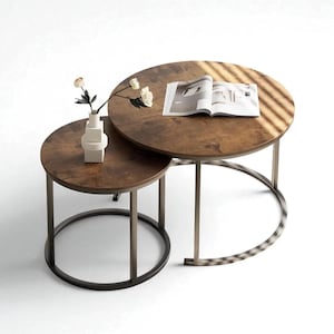 Peut inclure: Ensemble de deux tables basses gigognes. Elles présentent des plateaux ronds bruns avec une finition texturée et des cadres en métal foncé. Un vase avec des fleurs et un magazine ouvert sont sur les tables.