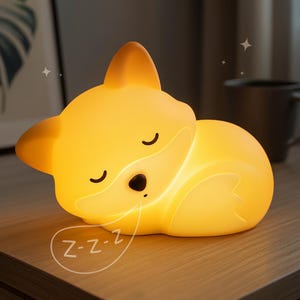 May include: A glowing yellow fox-shaped night light. The light is in the shape of a sleeping fox with closed eyes and a small black nose. A speech bubble with "Z-z-z" is next to the fox. The night light is on a wooden surface.