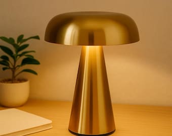 May include: A gold-colored table lamp with a mushroom-shaped shade and a conical base. The lamp is illuminated, casting a warm glow. The lamp is on a wooden surface, with a small potted plant and a notebook in the background.