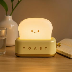 May include: A novelty night light shaped like a toaster with a slice of toast. The toast has a smiling face. The word "TOAST" is printed on the front of the toaster. The toaster is light yellow with gold accents.