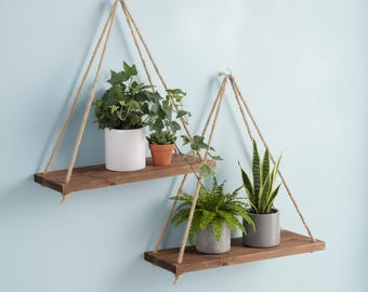 May include: Two hanging wooden shelves with plants. Each shelf is suspended by rope and holds potted greenery. The shelves are made of dark wood and are mounted against a light blue wall. The plants include ivy, succulents, and ferns.