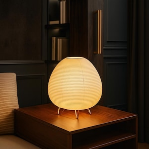 May include: A glowing, egg-shaped table lamp with a paper shade and a warm, inviting light. The lamp sits on a dark wooden table, supported by three slender, gold-colored legs. The lamp's design is minimalist and modern, suitable for a bedroom or living room.