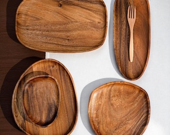 May include: A collection of wooden serving platters and a wooden fork. The platters are various shapes, including rectangular, oval, and round, with natural wood grain patterns. The fork is placed on an oval platter. The items are arranged on a white surface.
