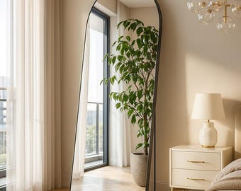 May include: A tall, arched-top mirror with a black frame. The mirror reflects a room with a large window, a potted plant, a nightstand, and a bed. The room has a neutral color palette with light-colored walls and curtains.