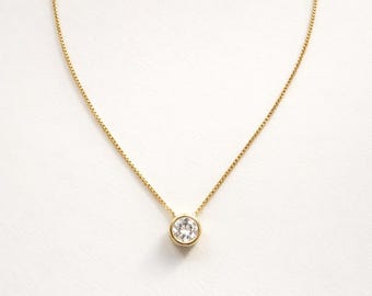 May include: A gold-colored necklace with a round, clear gemstone pendant. The pendant is set in a gold bezel and hangs from a delicate gold chain. The necklace is displayed against a white background.