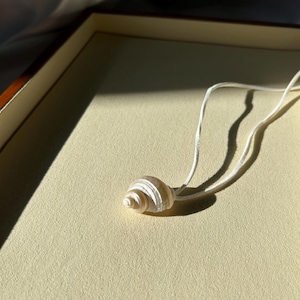 Puede incluir: Un delicado collar con colgante de concha. El colgante es una pequeña concha en espiral en tonos crema y blanco, suspendida de un fino cordón blanco. El collar descansa sobre una superficie amarillo claro, con sombras.