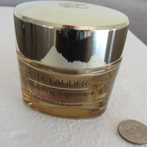 Könnte beinhalten: Ein Tiegel Estee Lauder Revitalizing Supreme+ Youth Power Creme. Der Tiegel ist transparent mit einem goldenen Deckel und goldfarbenen Buchstaben. Der Text "Estee Lauder Revitalizing Supreme+ Youth Power Creme" ist auf dem Tiegel aufgedruckt. Eine Münze liegt neben dem Tiegel.