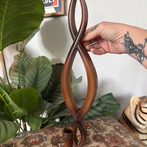 May include: A tall, brown, sculptural candle holder featuring an elongated, twisted infinity loop design that rises vertically from a single, tapered, cone-shaped base designed to securely hold a candle in an upright position.