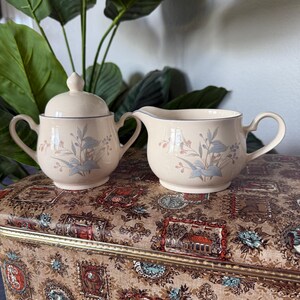 May include: A cream-colored sugar bowl with a lid and a creamer with a floral design. The set has blue and pink accents and a blue trim. The pieces are on a patterned surface.