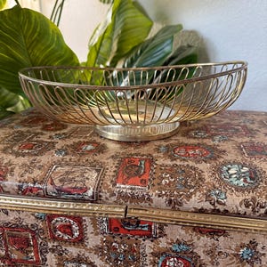 May include: An oval-shaped gold-toned wire basket bowl featuring a series of vertical metal bars forming the sides. The bowl has a solid circular base and an open top design, constructed from thin, polished metal wire with a reflective finish.