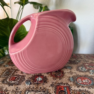 May include: A vintage pink ceramic pitcher with a large handle and a curved spout. The pitcher has a circular design on the body and is likely used for serving beverages. The pitcher is a soft pink color and is sitting on a patterned surface.