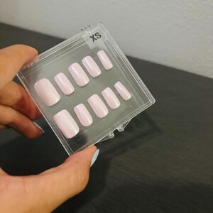 May include: A set of XS light pink press-on nails in a clear plastic case. The nails are various shapes and sizes, with a glossy finish. The case is held by a hand.