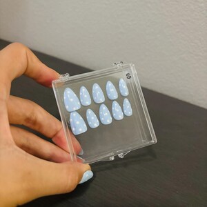 May include: A set of light blue press-on nails with white polka dots, displayed in a clear plastic case. The nails are almond-shaped and have a glossy finish. The case is labeled with the letter "S".