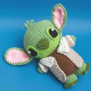 May include: A handmade crochet doll in the shape of a character with large ears and eyes. The doll is primarily green with pink accents on the ears and a brown and beige outfit. The doll is lying on a blue background.