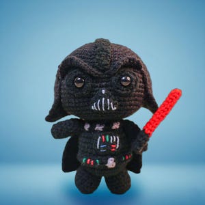 Könnte beinhalten: Gehäkelte Darth Vader Puppe in Schwarz mit rotem Lichtschwert. Die Puppe hat einen detaillierten Helm, Gesichtszüge und einen kleinen Umhang. Die Figur hat ein farbenfrohes Design auf Brust und Gürtel.