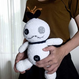 May include: A handmade white crochet doll with black stitched details, including an X on the belly and eyes. The doll has a gray eye and a black bow on top. The doll is being held by a person.
