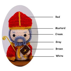 May include: A crocheted figure of a saint, featuring a red hat with a yellow stripe, a cream-colored face, and a gray beard. The figure holds a yellow staff and a brown book with a yellow cross. The figure is wearing a red robe.