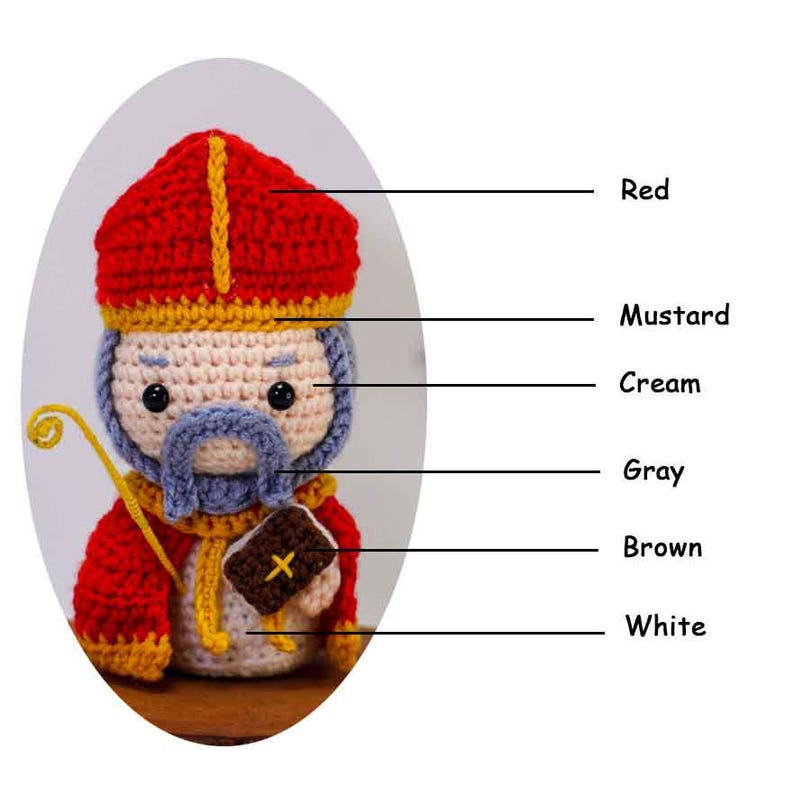 May include: A crocheted figure of a saint, featuring a red hat with a yellow stripe, a cream-colored face, and a gray beard. The figure holds a yellow staff and a brown book with a yellow cross. The figure is wearing a red robe.