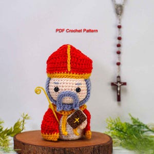 May include: A handmade crochet figure of a saint, featuring a red hat with yellow trim, a blue beard, and a staff. The figure is holding a small book. The text "PDF Crochet Pattern" is visible.