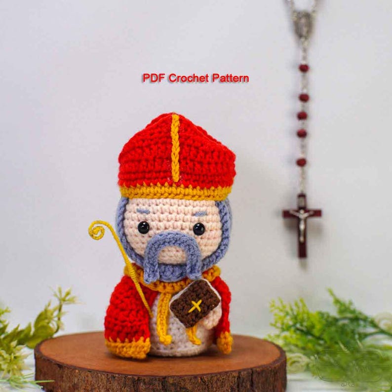 May include: A handmade crochet figure of a saint, featuring a red hat with yellow trim, a blue beard, and a staff. The figure is holding a small book. The text "PDF Crochet Pattern" is visible.