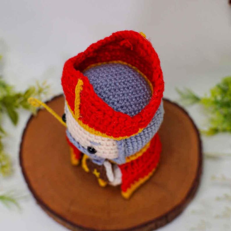 May include: Crocheted figure of a character wearing a red and yellow hat and robe. The figure has a gray head and a white face with black eyes. The figure is sitting on a round, brown wooden surface.