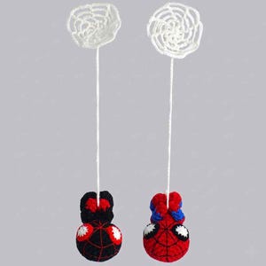 May include: Two crocheted figures hanging from white yarn strings attached to white circular web patterns. One figure is black with red web details and white eyes, while the other is red and blue with black web details and white eyes.
