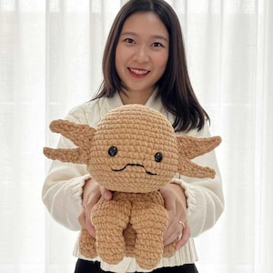 May include: A crocheted axolotl plushie in shades of tan and brown. The handmade toy features a rounded head, six frilly gills, and a smiling face with black button eyes. The plushie is held in front of a person.