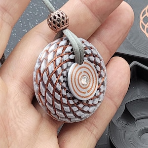 Op de afbeelding: Een handgemaakte hangerketting met een getextureerd, bolvormig ontwerp, koperdraad accenten en een kleine, cirkelvormige bedel met een spiraalpatroon. De hanger hangt aan een grijs koord met een decoratieve kraal.