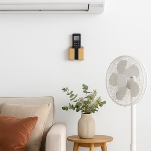 May include: A modern living room scene featuring a white air conditioner, a wooden remote control holder, and a standing fan. A beige sofa with brown and beige pillows sits near a small wooden side table with a vase of greenery.