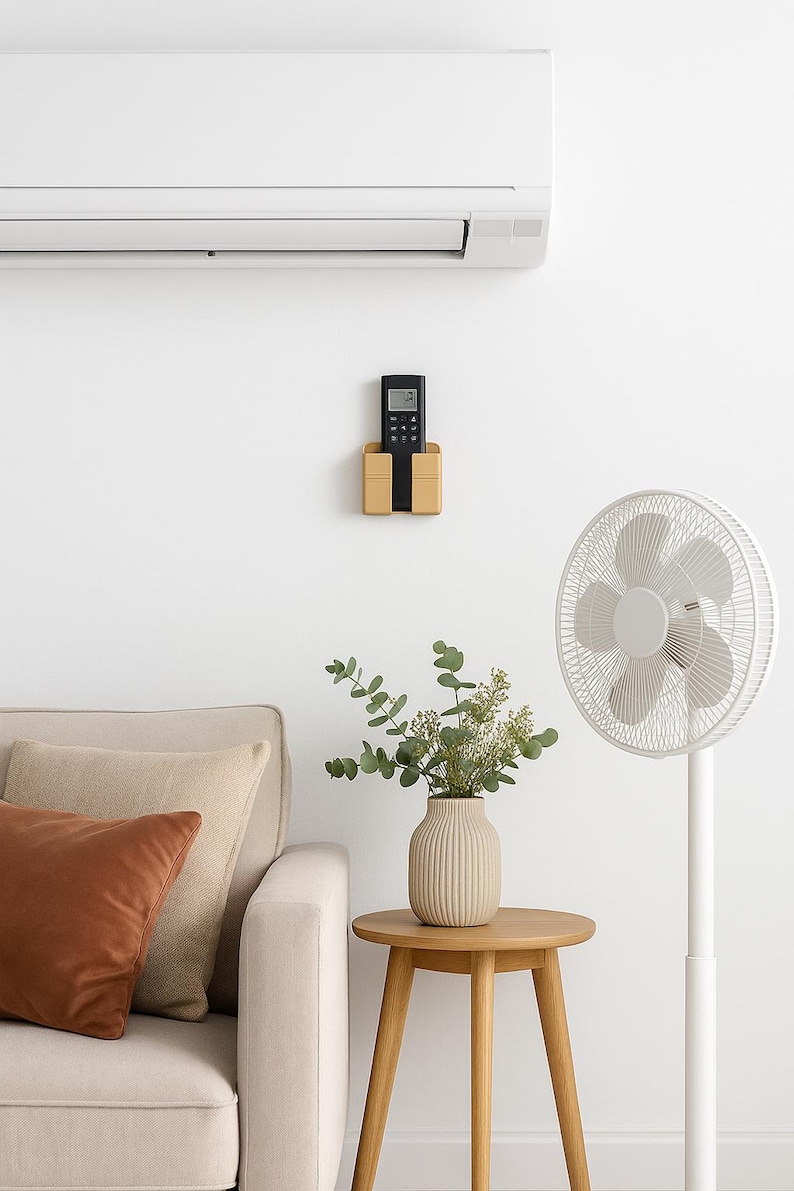 May include: A modern living room scene featuring a white air conditioner, a wooden remote control holder, and a standing fan. A beige sofa with brown and beige pillows sits near a small wooden side table with a vase of greenery.