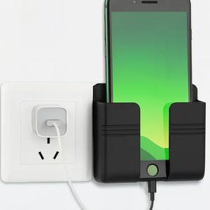 May include: A black phone holder with a smartphone charging. The phone has a green screen. A white electrical outlet is to the left. The phone holder is mounted on a white wall.