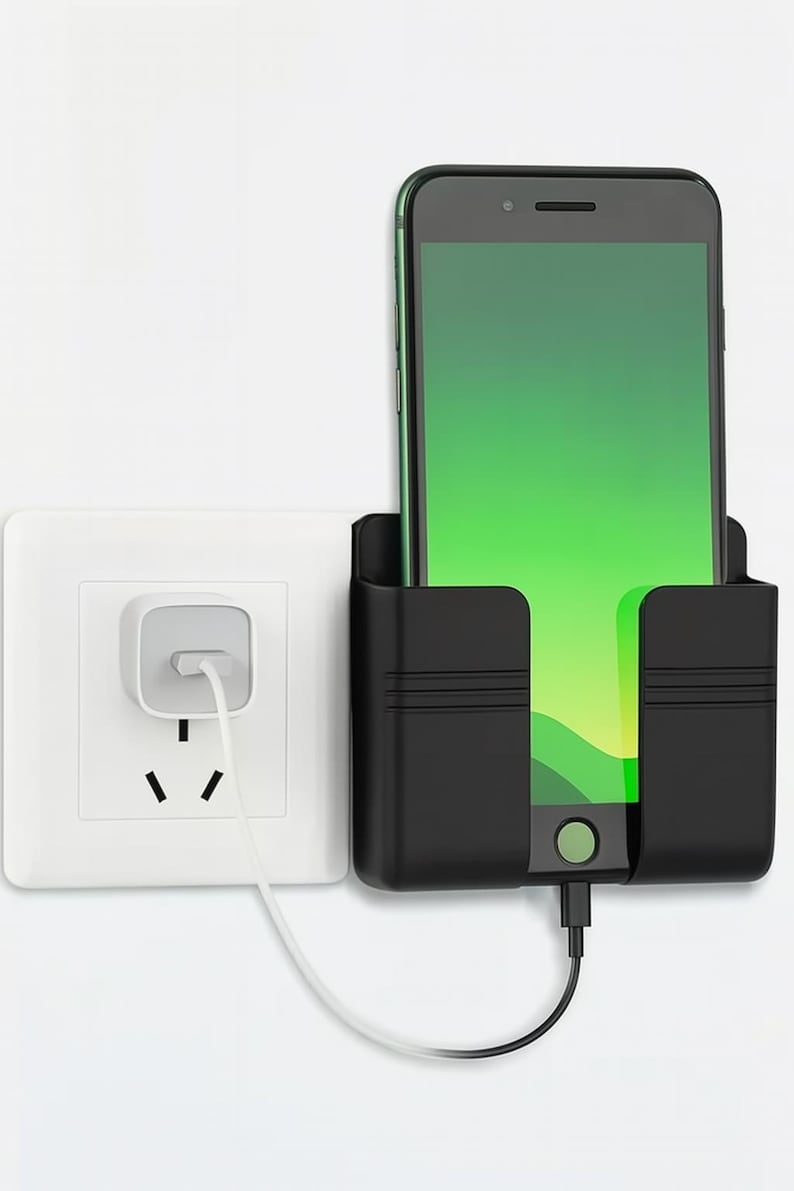 May include: A black phone holder with a smartphone charging. The phone has a green screen. A white electrical outlet is to the left. The phone holder is mounted on a white wall.