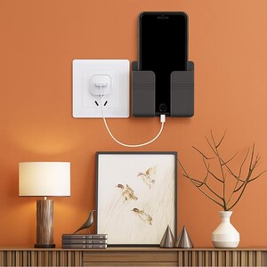 May include: A black smartphone is placed in a wall-mounted charging station, connected to a white electrical outlet via a charging cable. The setup is against an orange wall, with a framed artwork and decorative items on a wooden surface below.