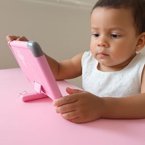 K&ouml;nnte beinhalten: Ein junges Kind sitzt an einem rosa Tisch und h&auml;lt ein rosa Tablet. Das Tablet hat einen St&auml;nder und einen grauen Akzent an der oberen Ecke. Das Kind tr&auml;gt ein wei&szlig;es &auml;rmelloses Kleid und schaut auf das Tablet.