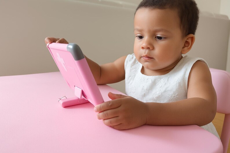 K&ouml;nnte beinhalten: Ein junges Kind sitzt an einem rosa Tisch und h&auml;lt ein rosa Tablet. Das Tablet hat einen St&auml;nder und einen grauen Akzent an der oberen Ecke. Das Kind tr&auml;gt ein wei&szlig;es &auml;rmelloses Kleid und schaut auf das Tablet.