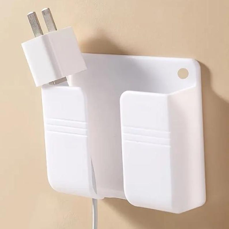 May include: A white plastic wall-mounted phone charging station with two compartments and a plug inserted. The station has a rectangular back with a hole for hanging. The design is simple and functional, ideal for organizing charging cables.