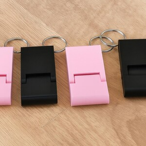 K&ouml;nnte beinhalten: Vier Schl&uuml;sselanh&auml;nger mit Handy-St&auml;nder-Funktion. Zwei sind hellrosa und zwei sind schwarz. Jeder hat einen rechteckigen K&ouml;rper mit einem klappbaren St&auml;nder und einem silbernen Schl&uuml;sselring. Die Artikel liegen auf einer hellbraunen Holzoberfl&auml;che.