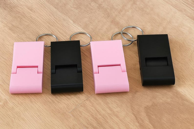 K&ouml;nnte beinhalten: Vier Schl&uuml;sselanh&auml;nger mit Handy-St&auml;nder-Funktion. Zwei sind hellrosa und zwei sind schwarz. Jeder hat einen rechteckigen K&ouml;rper mit einem klappbaren St&auml;nder und einem silbernen Schl&uuml;sselring. Die Artikel liegen auf einer hellbraunen Holzoberfl&auml;che.