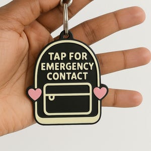 Puede incluir: Llavero en blanco y negro con el texto "TAP FOR EMERGENCY CONTACT". El llavero tiene un diseño de mochila con detalles de corazón rosa. Está sujeto a un cierre de metal plateado.