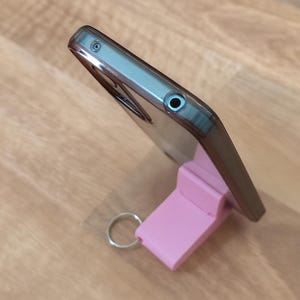 K&ouml;nnte beinhalten: Ein Smartphone in einer transparenten H&uuml;lle steht auf einem rosa St&auml;nder mit einem silbernen Schl&uuml;sselring. Das Telefon ist abgewinkelt und zeigt sein Profil. Der St&auml;nder hat eine rechteckige Form.