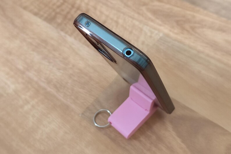 K&ouml;nnte beinhalten: Ein Smartphone in einer transparenten H&uuml;lle steht auf einem rosa St&auml;nder mit einem silbernen Schl&uuml;sselring. Das Telefon ist abgewinkelt und zeigt sein Profil. Der St&auml;nder hat eine rechteckige Form.