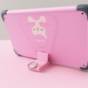 K&ouml;nnte beinhalten: Rosa Tablet-H&uuml;lle mit integriertem St&auml;nder und einem Cartoon-Hasen-Motiv. Die H&uuml;lle hat graue Ecksch&uuml;tzer und den Text "WeeKidz". Der St&auml;nder hat einen silbernen Ring.