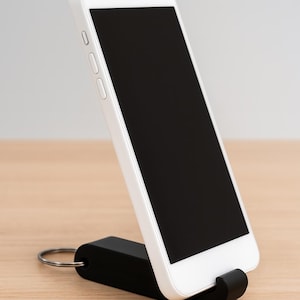 May include: A white smartphone with a black screen is held by a black phone stand. The stand has a small loop for a keyring. The phone is angled, showing its screen.